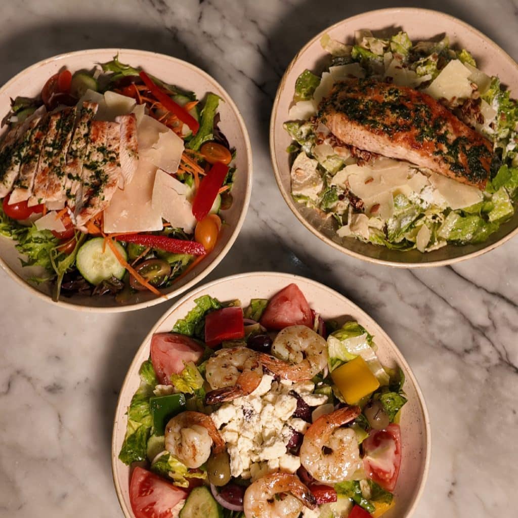 Three bowls of colorful salads on a marble table: grilled chicken with greens and shaved parmesan; a second chicken salad; and a shrimp, feta, and veggie salad.