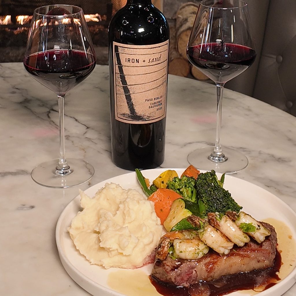 Plate of steak topped with shrimp, mashed potatoes, and roasted vegetables on a marble table with a bottle of Iron + Sand wine and two red-wine glasses by a lit fireplace.