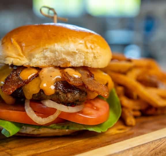Juicy cheeseburger with bacon, melted cheese, tomato, onion slices and lettuce on a toasted bun, with fries on a wooden board behind it