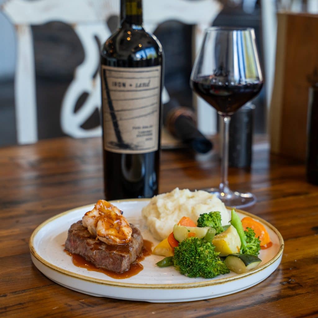 Plate dinner featuring a steak topped with shrimp, mashed potatoes and mixed vegetables; a wine bottle and glass appear in the background.