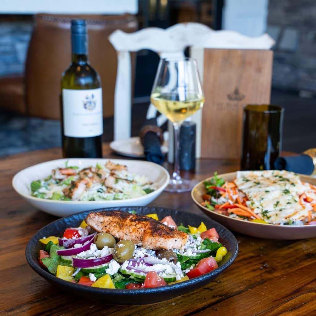 Two large bowls of colorful salad with greens, tomatoes, peppers, olives, and feta, plus a grilled chicken breast on a dark plate in the foreground, with a bottle of white wine and glass in the background.