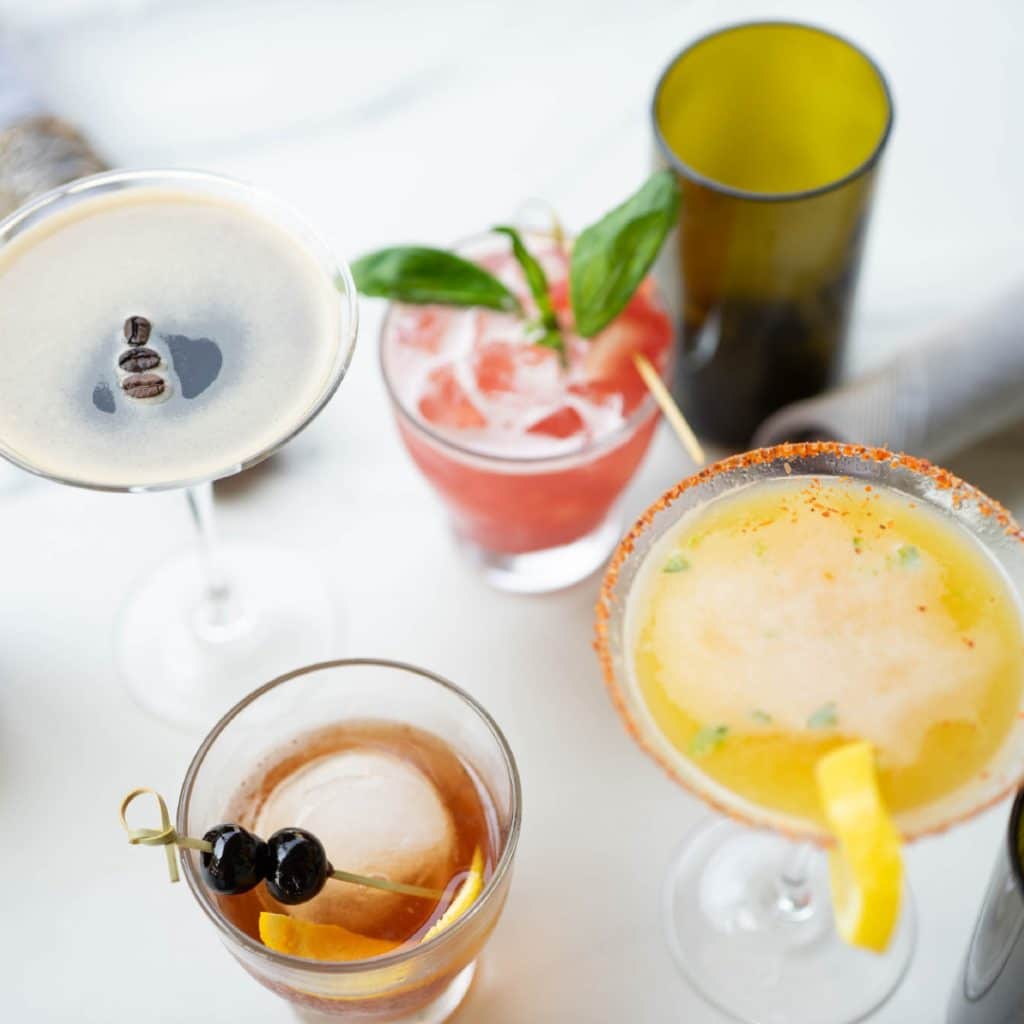 Assorted cocktails on a white table: a frothy martini with coffee beans, a pink basil cocktail, a salted margarita, a citrus drink with lemon twist, and an old-fashioned-style drink with an orange garnish.