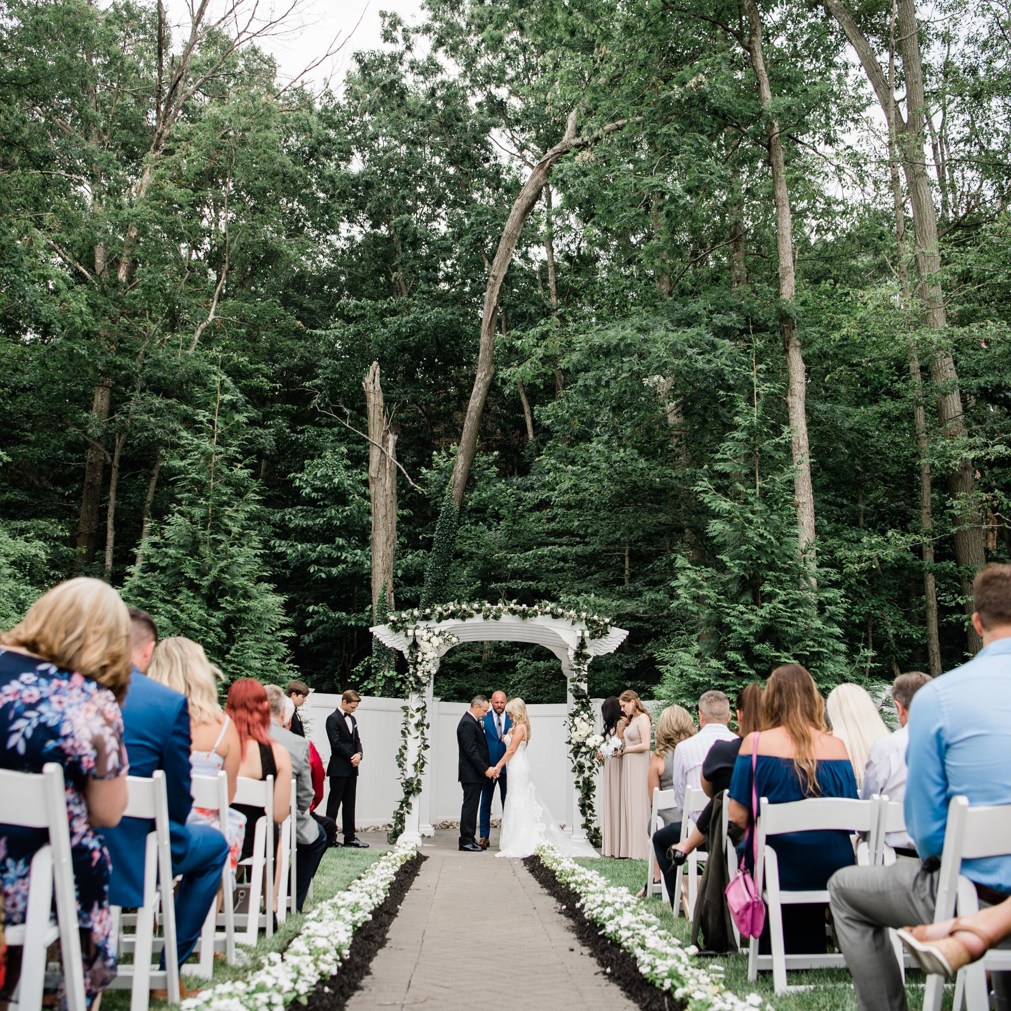 Bride and groom stand under a white floral arch exchanging vows as guests sit on either side outdoors.