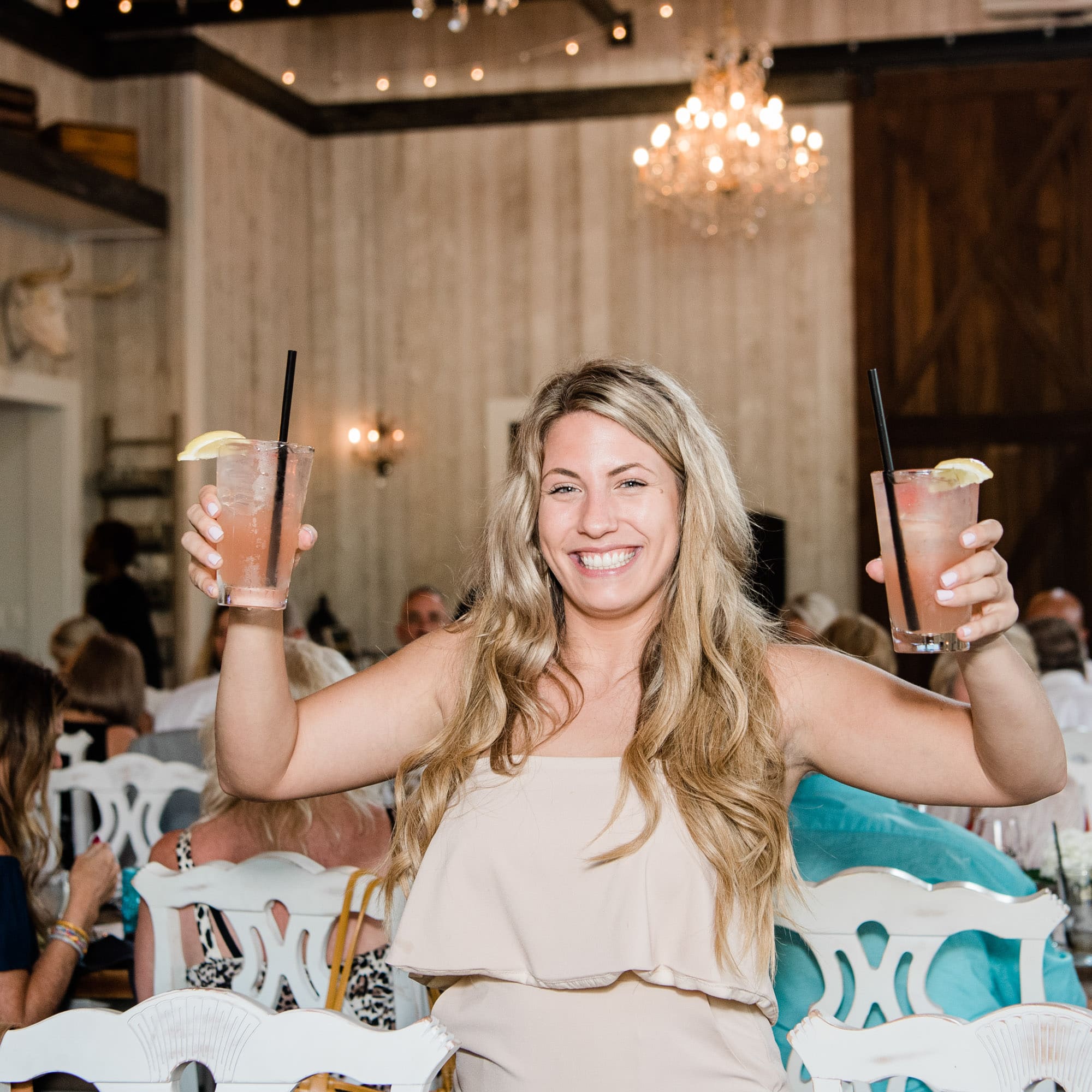 Smiling woman toasts with two pink cocktails, lemon wedges, at a festive party with guests and chandeliers in the background.