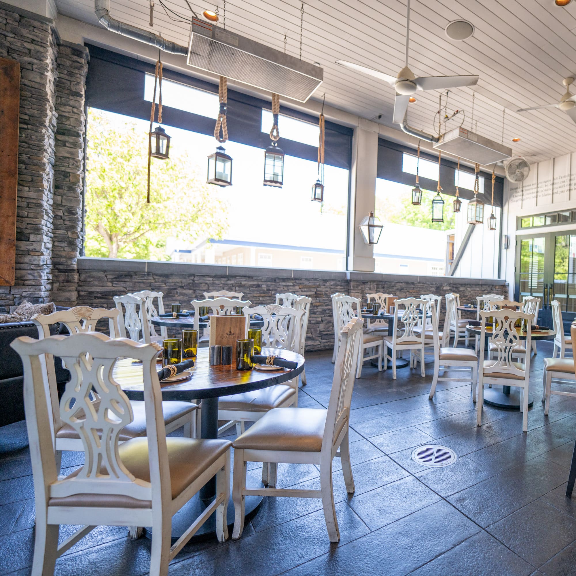 Indoor cafe dining area with white ornate chairs around round tables, stone wall backdrop, and hanging lantern decorations.