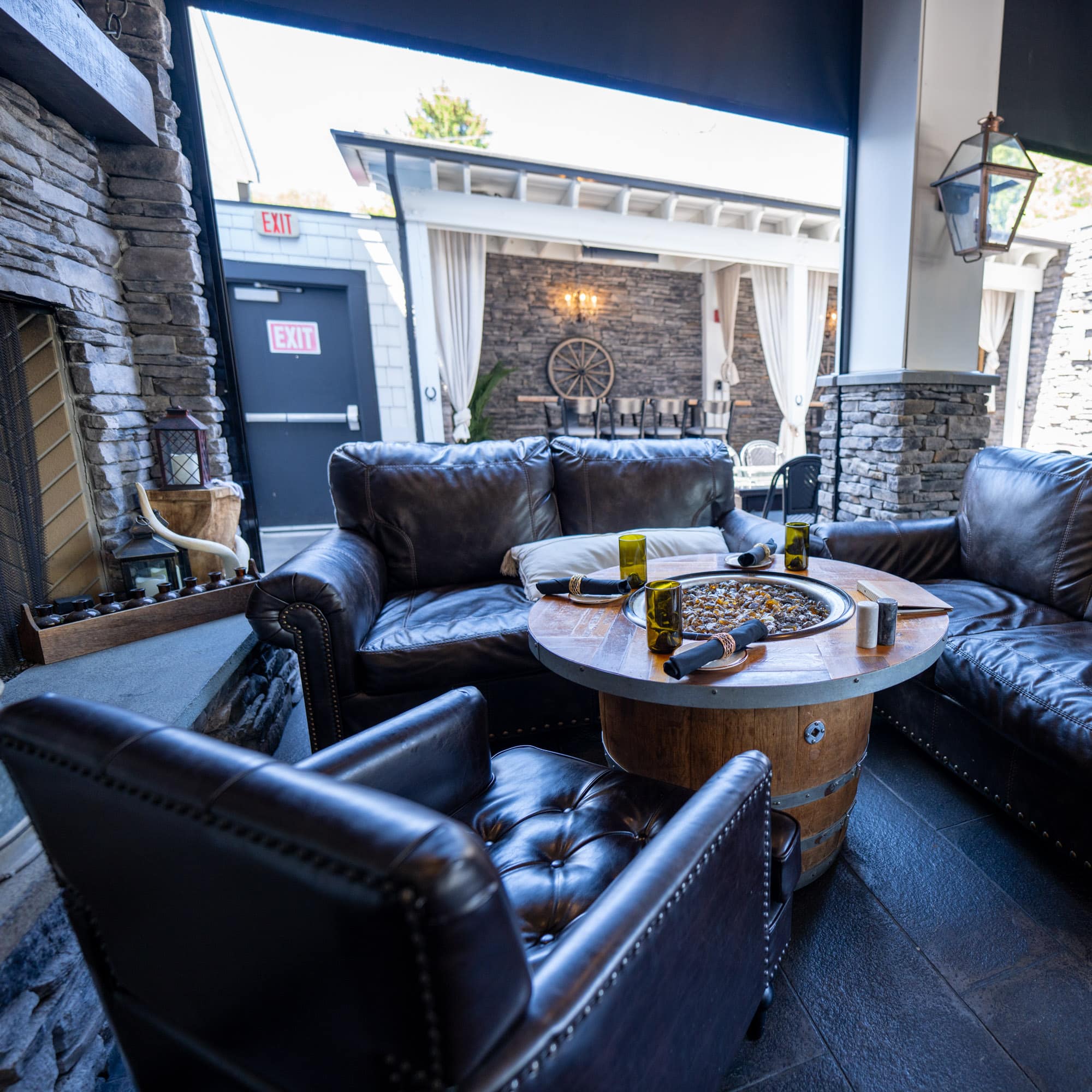 Outdoor lounge area with dark leather sectional sofas arranged around a round fire pit, stone walls and lanterns nearby.