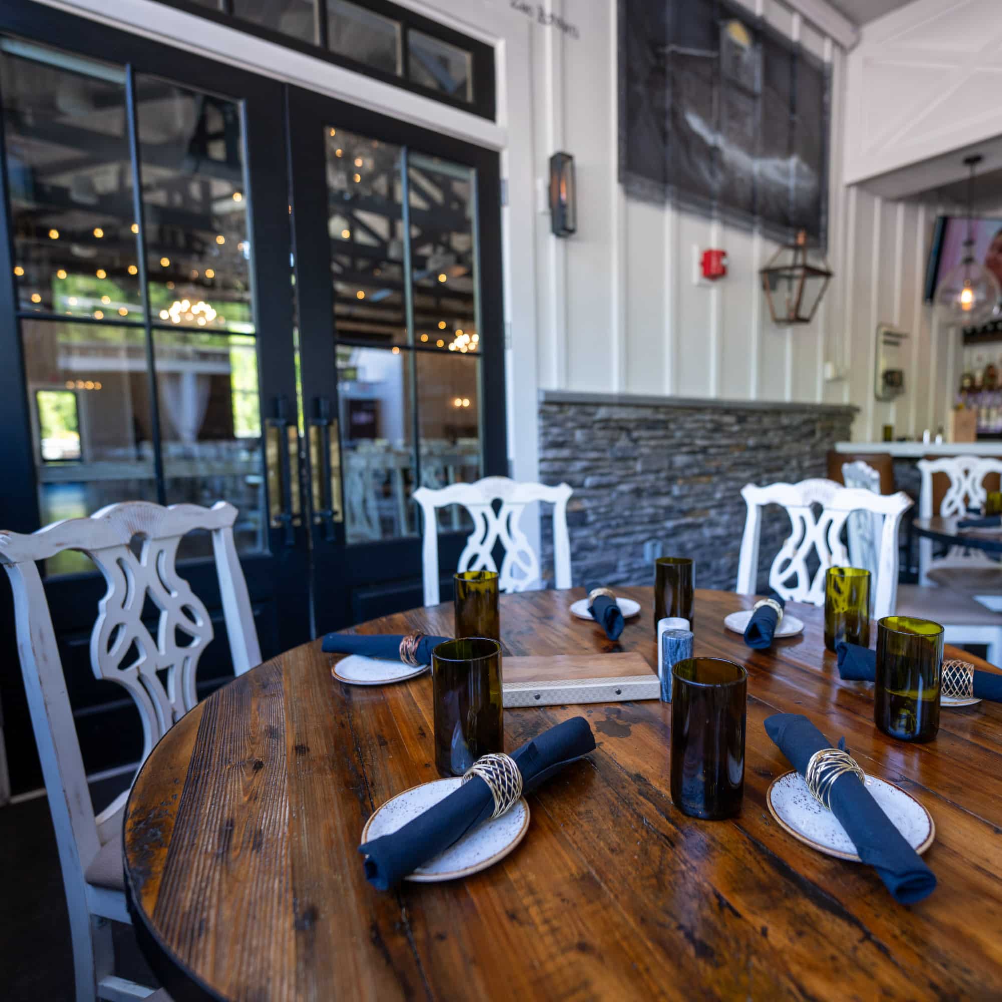 A round wooden table set with plates, navy napkins, and brown glass candle holders in a bright dining area with white chairs and glass doors behind.
