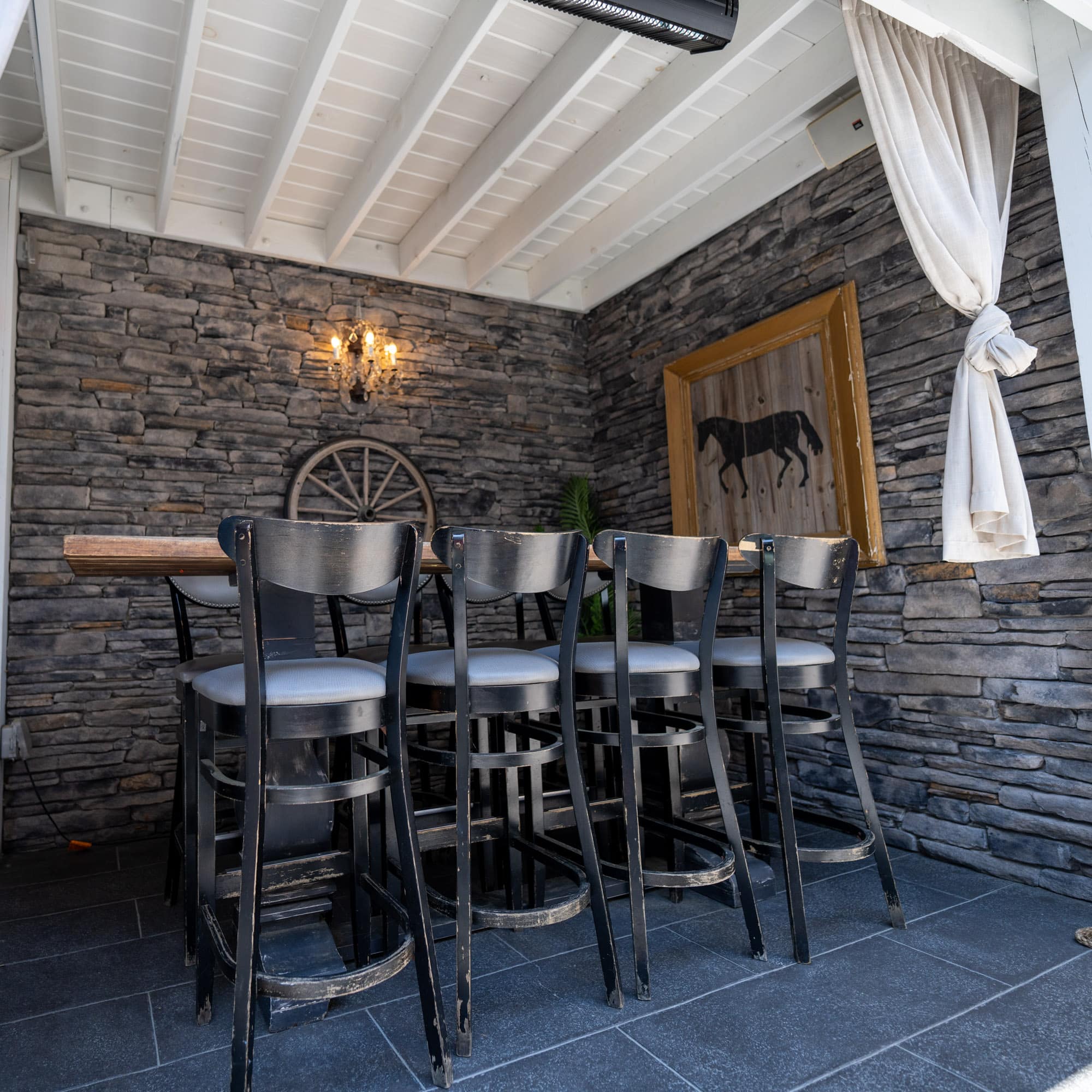 Row of black metal bar stools with light cushions along a wooden counter, set against a stacked stone wall with a chandelier and white ceiling beams in a cozy bar area.