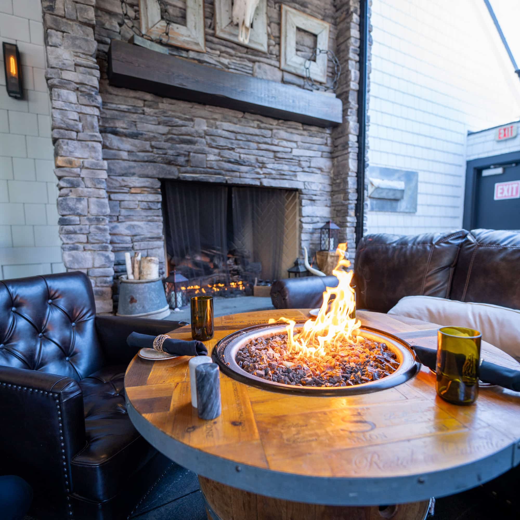 Outdoor seating area with a lit fire pit, surrounding black leather couches, and a stone wall backdrop, indoors/outdoors setting visible behind.