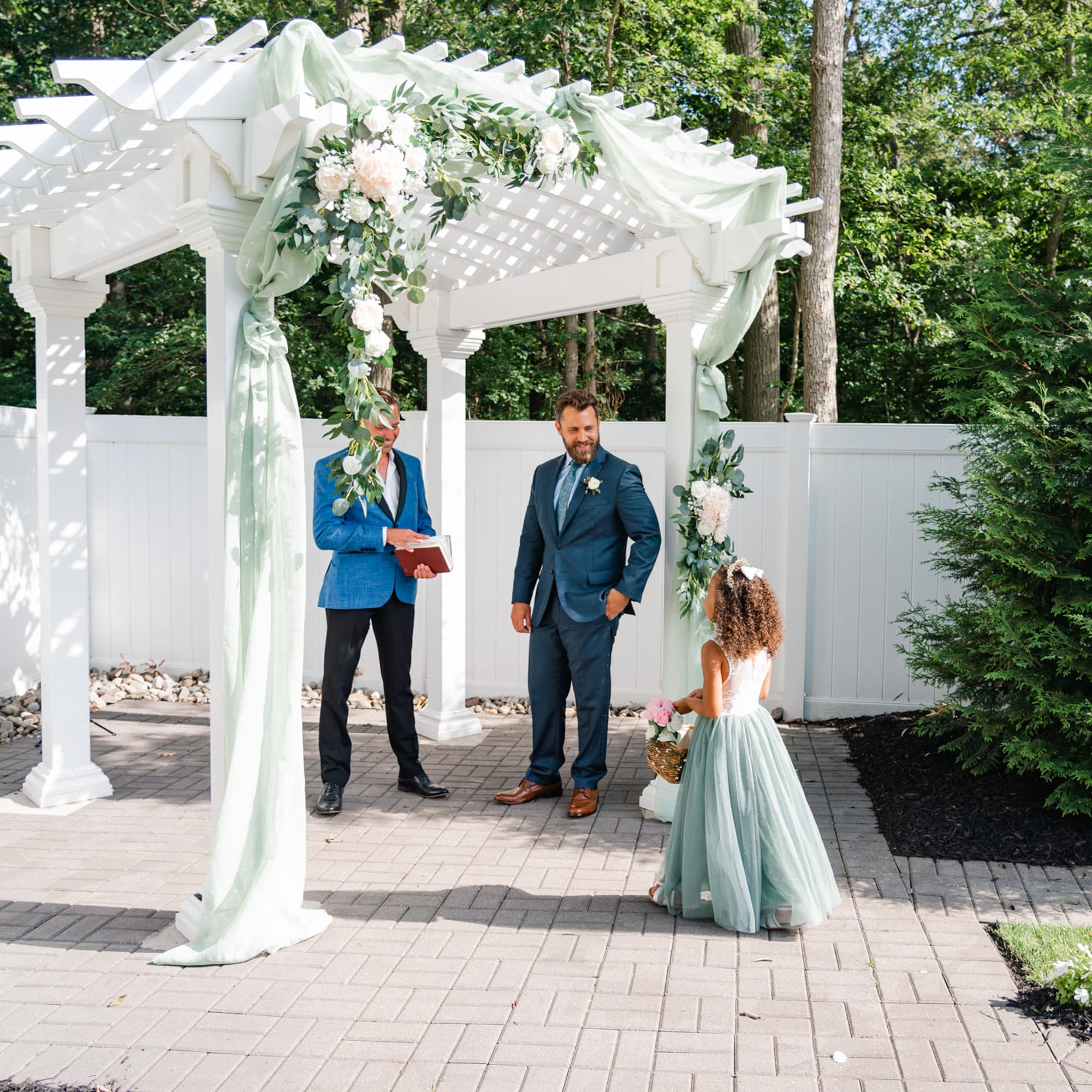 Outdoor wedding ceremony under a white pergola with a floral greenery arch; two men in suits stand nearby, a flower girl in a mint dress holds a basket.