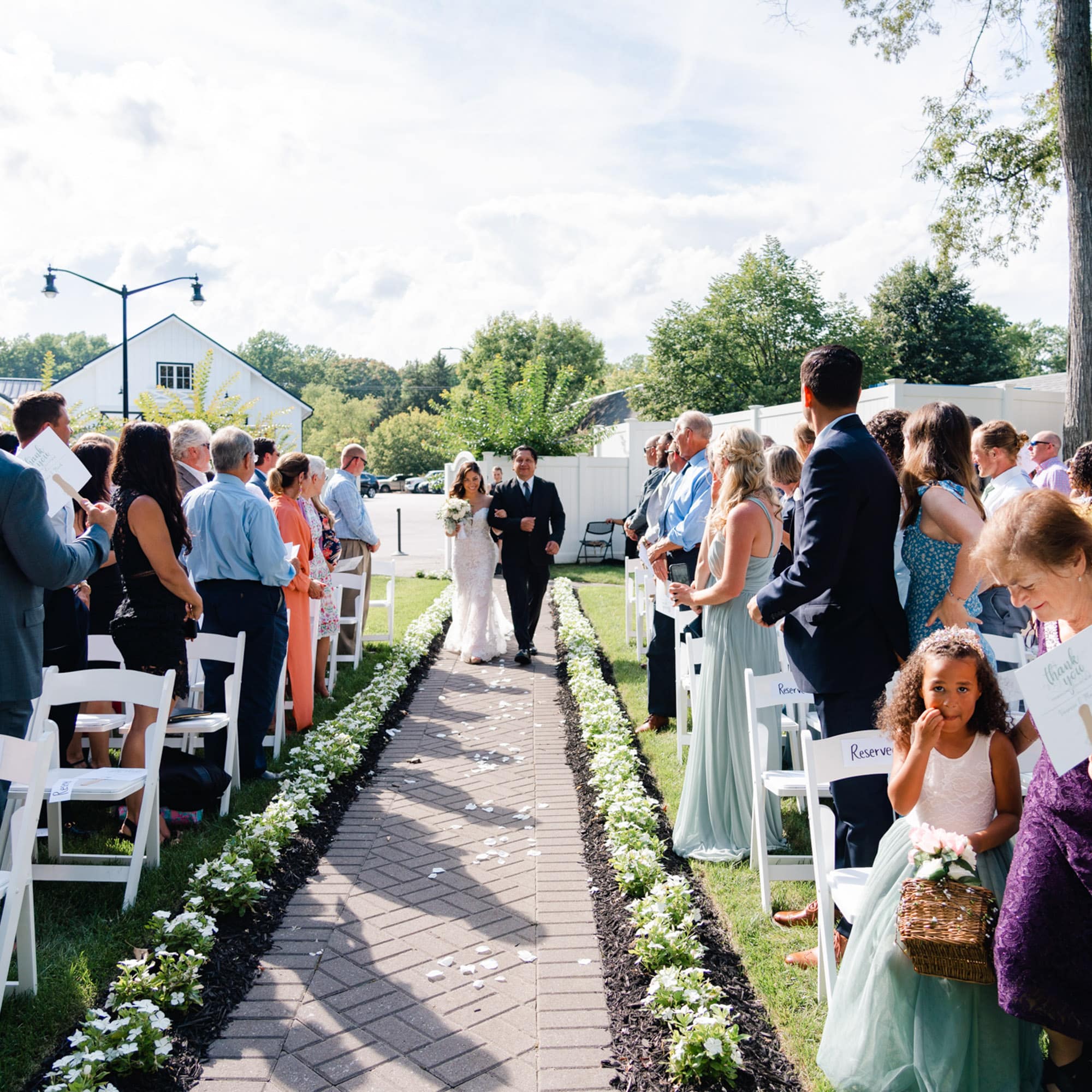 Bride in a white lace gown walking down the aisle with a man in a suit, escorted by guests standing on both sides in an outdoor ceremony.