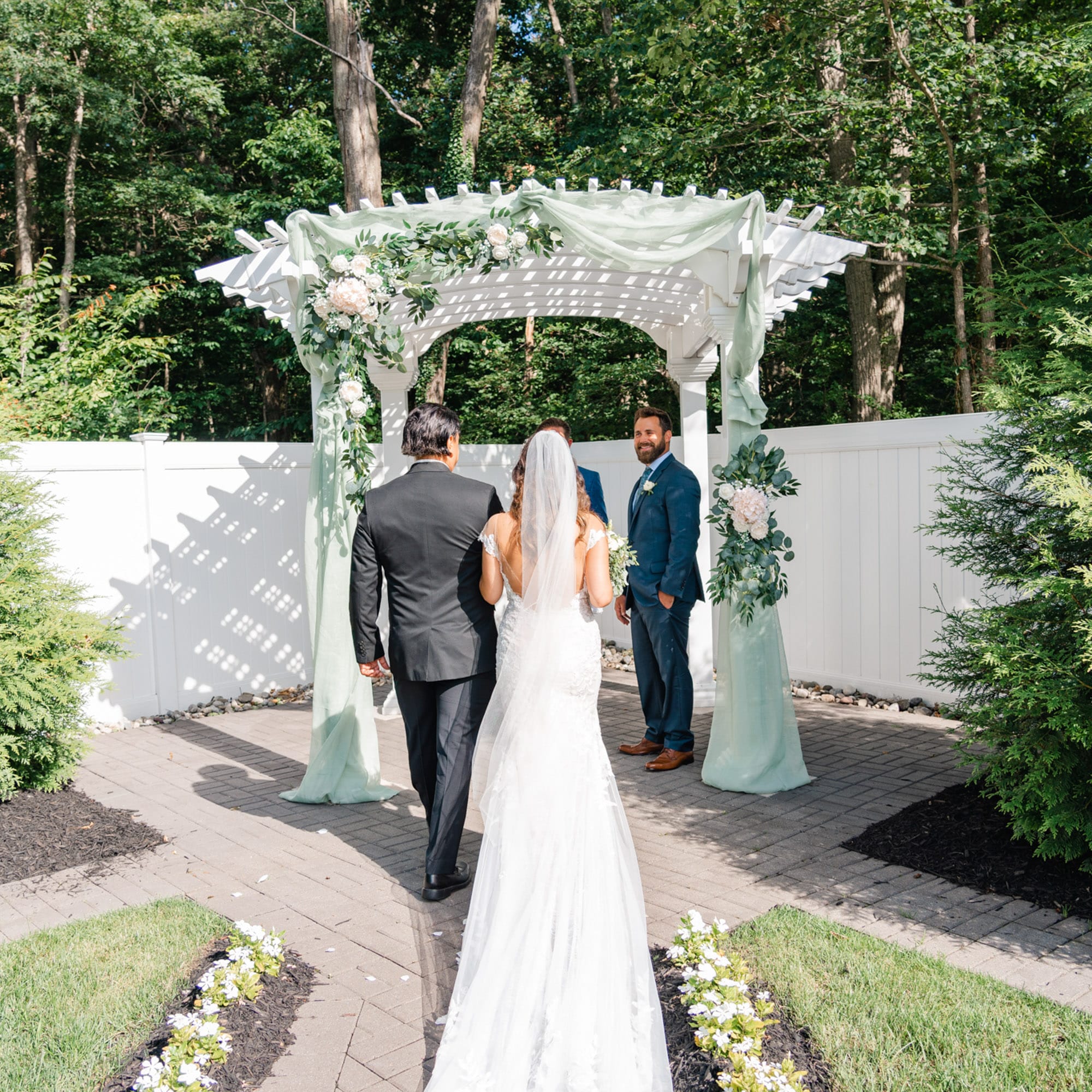 Bride in a white lace wedding gown and veil stands with the groom in a blue suit under a white arbor adorned with greenery and pale flowers in an outdoor garden setting.