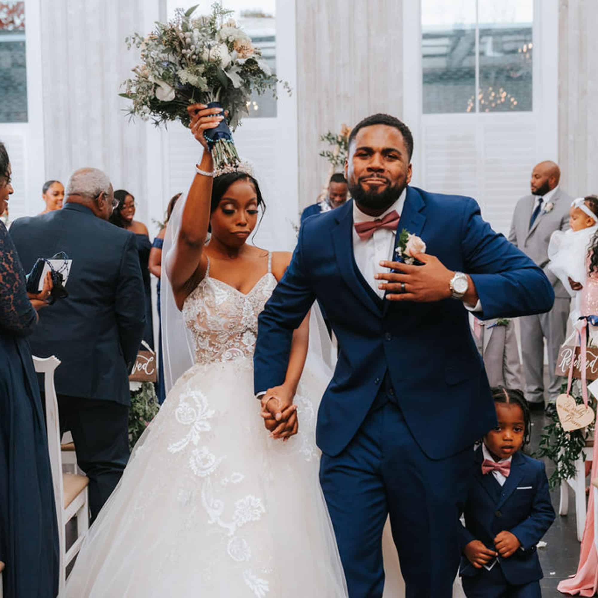Bride and groom walk together at a wedding reception, holding hands while guests cheer; the bride raises a bouquet aloft.