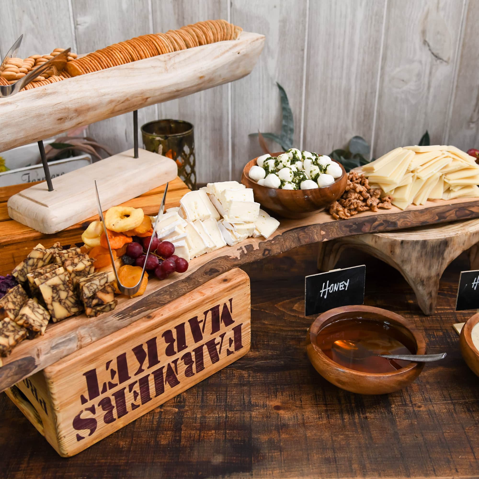 Rustic cheese platter on a wooden board with sliced cheeses, grapes, nuts, and a bowl of herb-coated mozzarella balls; honey sign in background.