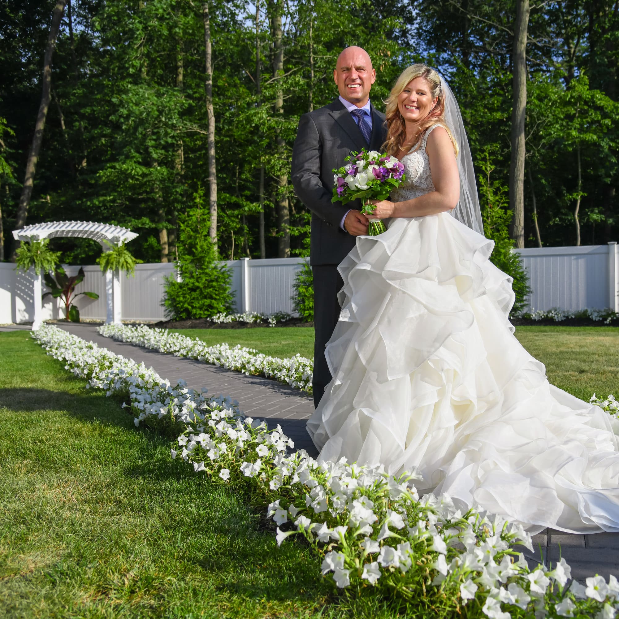 A newlywed couple stands outdoors; bride in a white gown holding a bouquet of white and purple flowers, groom in a dark suit smile together against a green garden backdrop.