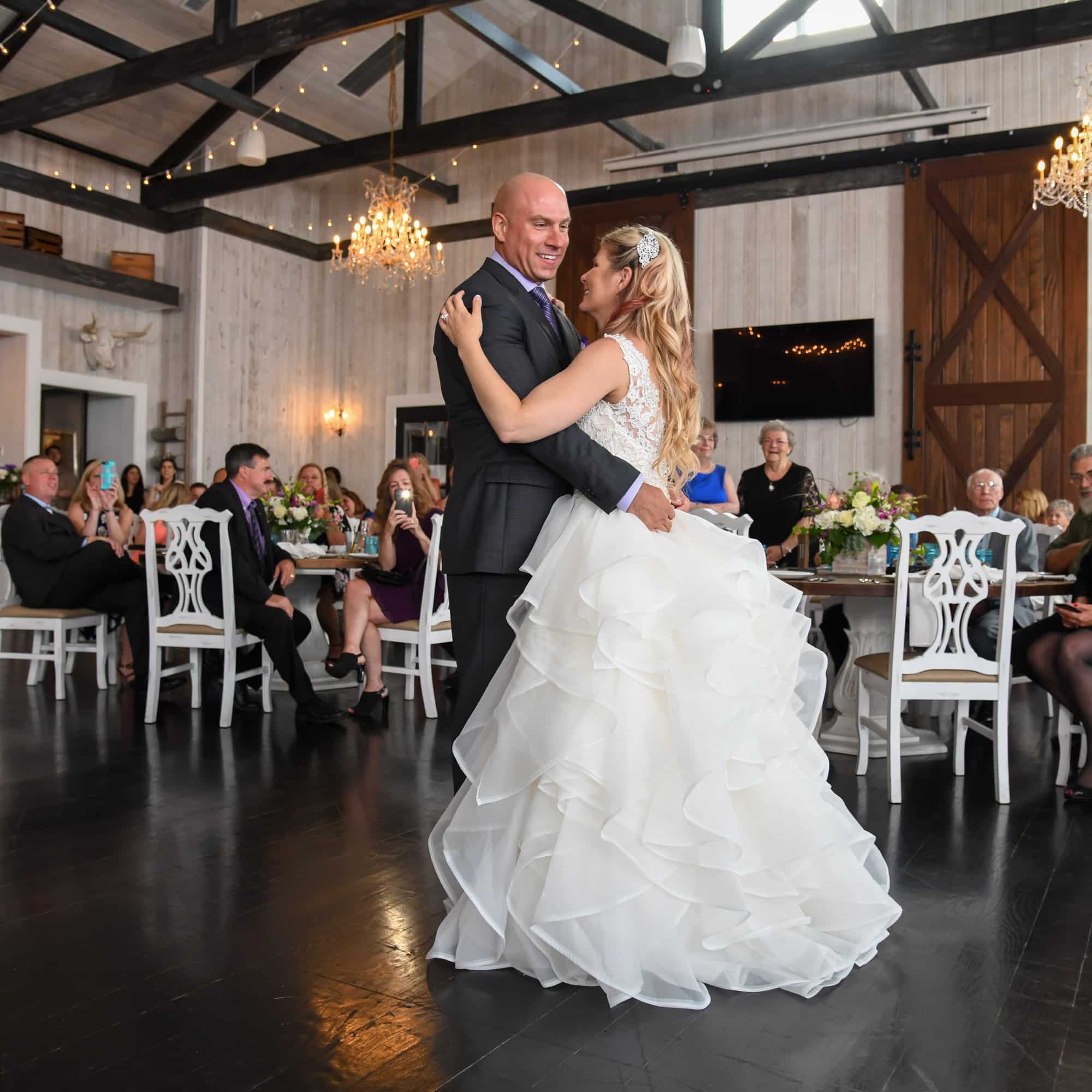 Bride in a white layered wedding gown dancing with a man in a dark suit at a wedding reception, guests seated at tables in a rustic, chandelier-lit venue behind them.