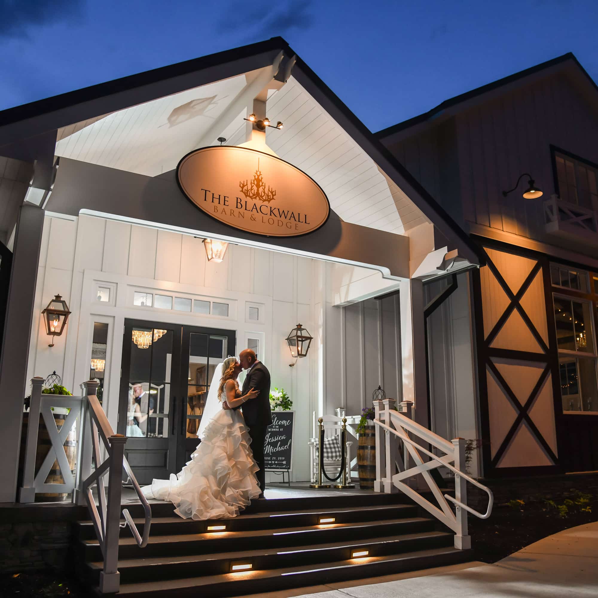 Bride and groom share a kiss on the lit venue steps, with a white storefront and the circular 'The Blackwell Barn & Lodge' sign above.