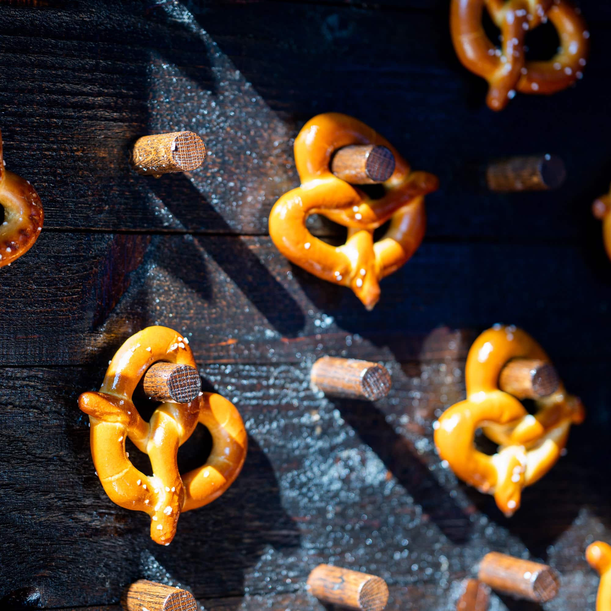 Close-up of glossy glazed pretzels scattered on a dark wooden surface with a few cinnamon sticks and salt crystals nearby.