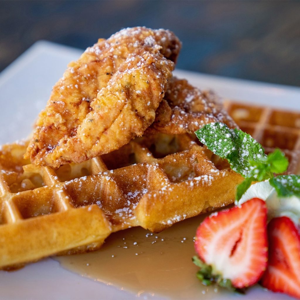 Waffle with a crispy fried chicken strip on top, powdered sugar, syrup, and a strawberry-mint garnish on the side.