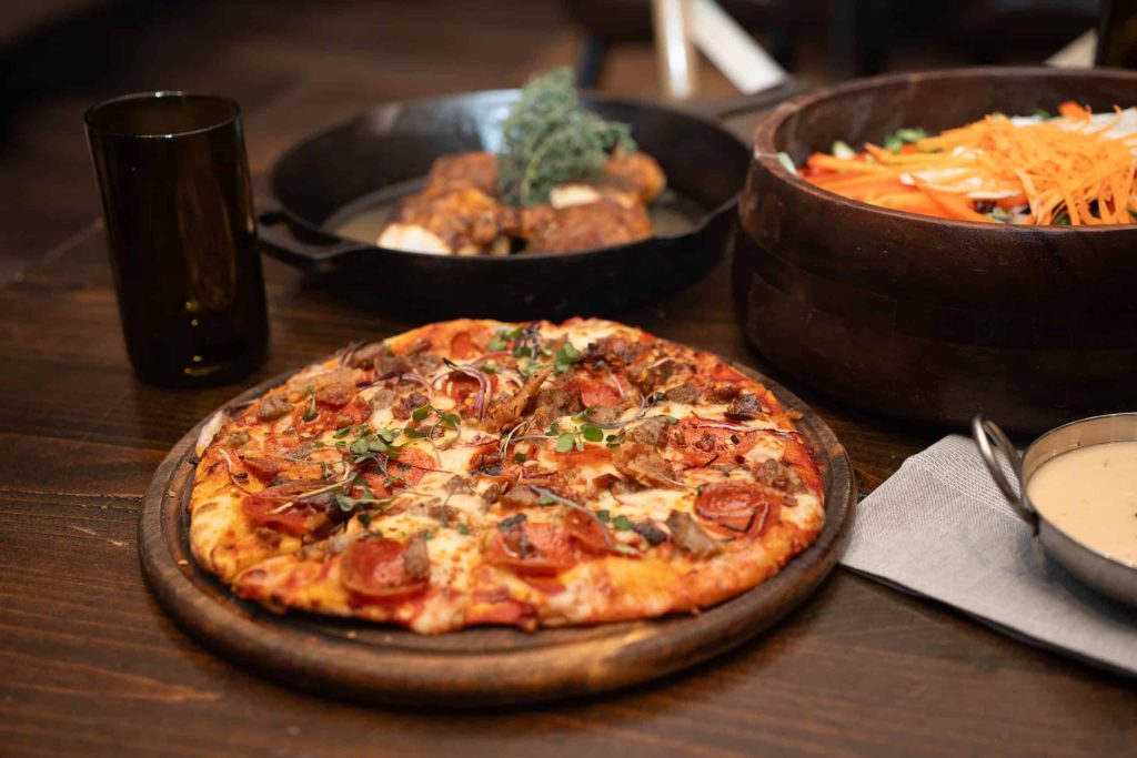 Pizza on a wooden board topped with pepperoni and herbs, with a dark glass and bowls in the background.