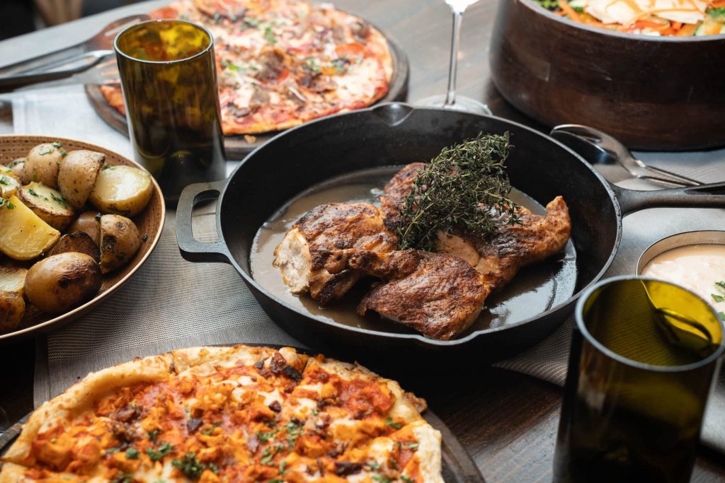 Shared table with a roast in a skillet topped with thyme, roasted potatoes, pizza, and bowls of salad and dipping sauces nearby.