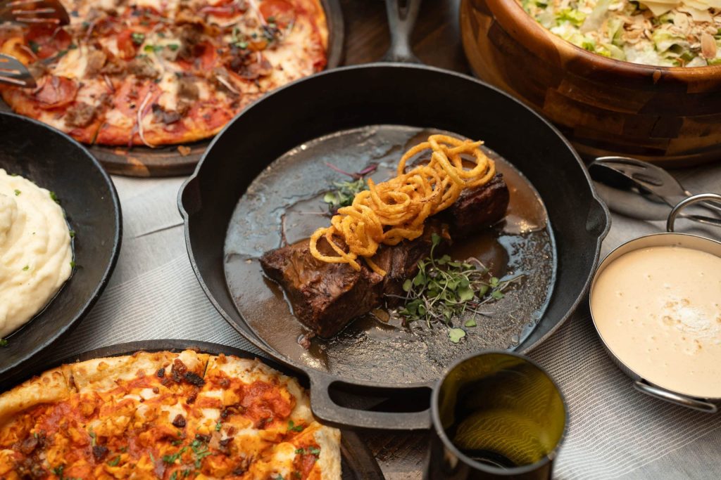 Beef in a black skillet topped with fried onion strings, surrounded by pizza slices and dips on a table.