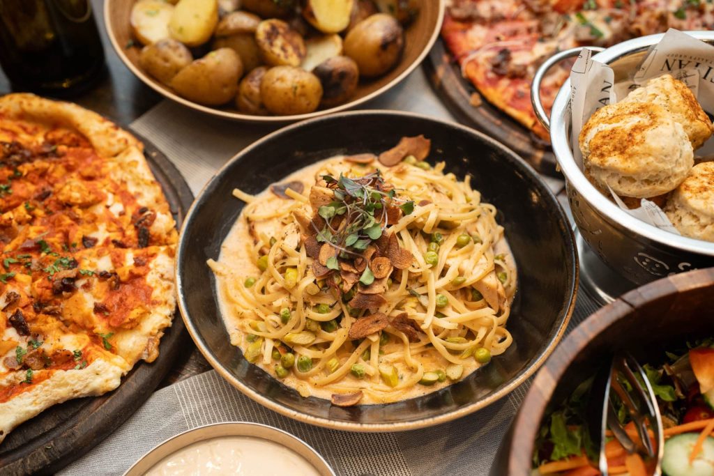 Bowl of pasta with peas and sliced mushrooms, topped with microgreens, surrounded by bread and pizzas on a rustic table spread