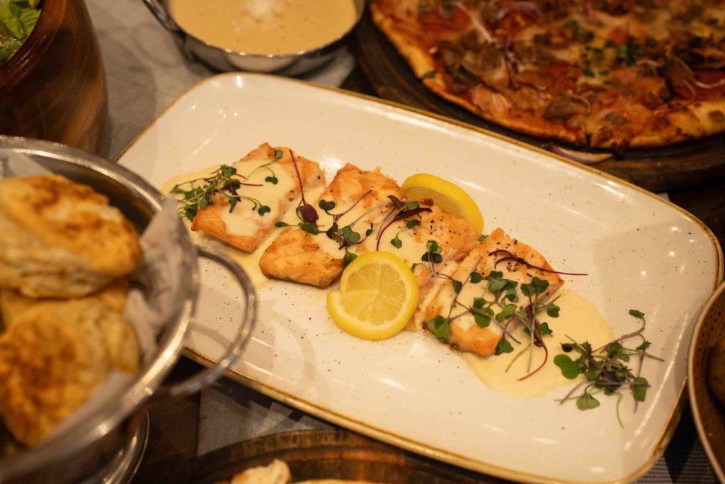 Plate of pan-seared fish topped with microgreens and lemon wedges, drizzled with a creamy sauce on a speckled beige plate; pizza visible in the background.